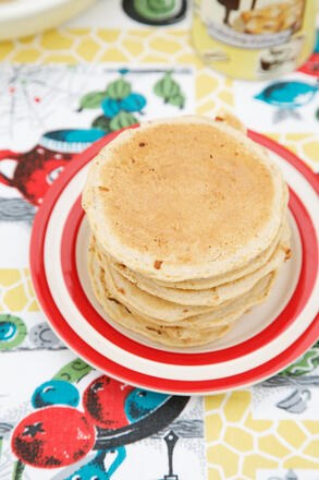 Pile of small pancakes stacked on red rimmed plate on vintage tablecloth with Honeybuns cake mix in the backgorund