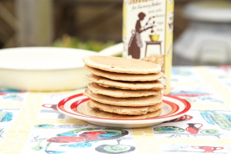 Pile of small pancakes stacked on red rimmed plate on vintage tablecloth with gluten free cake mix in the backgorund